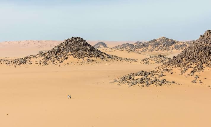 Open sand and dark rock outcrops in the desert, with two people walking in the distance.